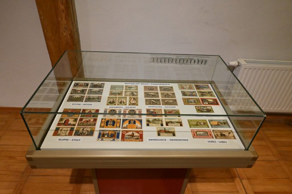 Gablota z zestawem historycznych banknotów i materiałów papierowych; na dużej planszy rozmieszczono liczne banknoty o różnych kolorach, formatach i wzorach, a poszczególne grupy zostały opisane i uporządkowane tematycznie (kliknięcie powiększy zdjęcie).