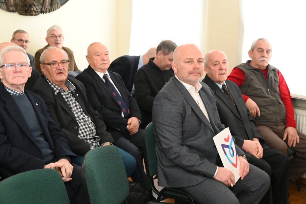 Grupa uczestników uroczystości siedzących obok siebie w pierwszym rzędzie; osoby pozują do wspólnego zdjęcia; w tle widoczni są pozostali goście spotkania (kliknięcie powiększy zdjęcie).
