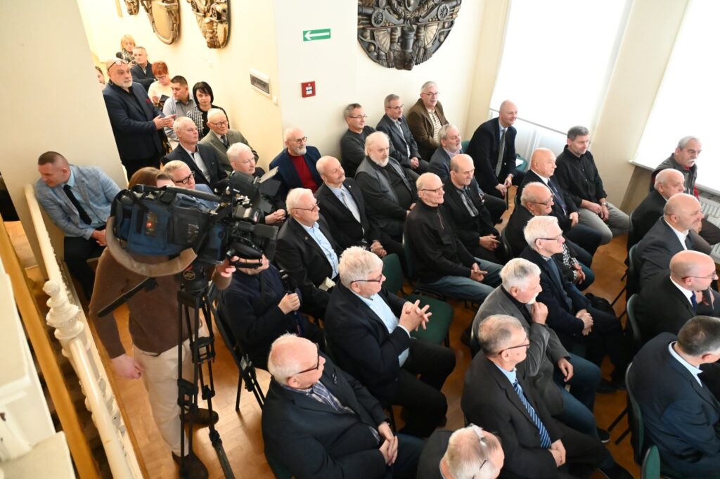 Uczestnicy uroczystego otwarcia wystawy zgromadzeni w sali; publiczność siedzi na krzesłach i obserwuje przebieg wydarzenia, a wśród obecnych znajduje się ekipa nagrywająca spotkanie; na ścianach widoczne są elementy wystroju muzeum (kliknięcie powiększy zdjęcie).