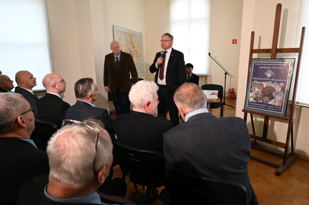 Fragment uroczystości związanej z otwarciem wystawy; przybyli goście siedzą na widowni i słuchają przemówienia osoby stojącej przy mikrofonie; obok znajduje się plansza informacyjna dotycząca wydarzenia ustawiona na sztaludze (kliknięcie powiększy zdjęcie).