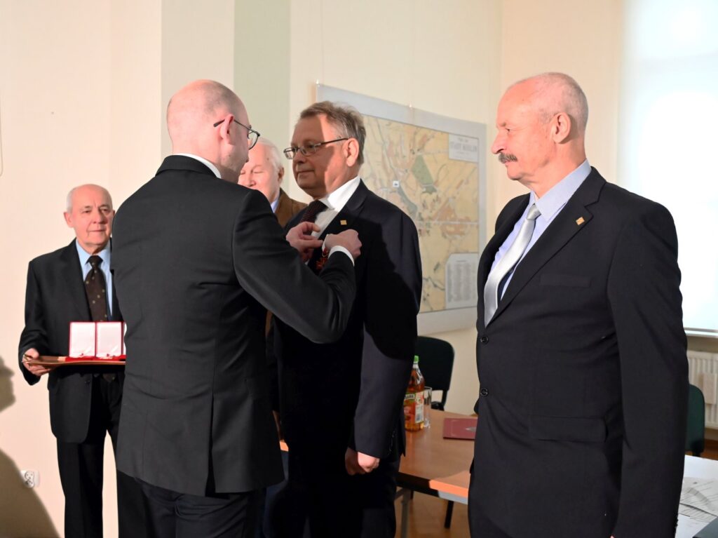 Moment uroczystego wręczania wyróżnienia podczas otwarcia wystawy; przedstawiciel organizatorów przypina odznaczenie jednemu z uczestników, a obok stoją kolejne osoby zaangażowane w wydarzenie (kliknięcie powiększy zdjęcie).