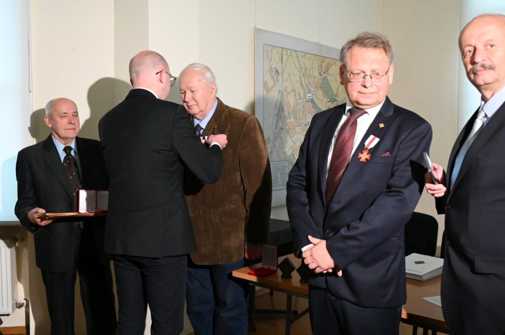 Uroczyste wręczenie odznaczenia podczas otwarcia wystawy; w centrum fotografii przedstawiciel organizatorów przypina wyróżnienie uczestnikowi wydarzenia, a obok stoją pozostali goście; jedna z osób trzyma otwarte etui z odznaczeniem, a uczestnik w pierwszym planie prezentuje przypięty medal (kliknięcie powiększy zdjęcie).