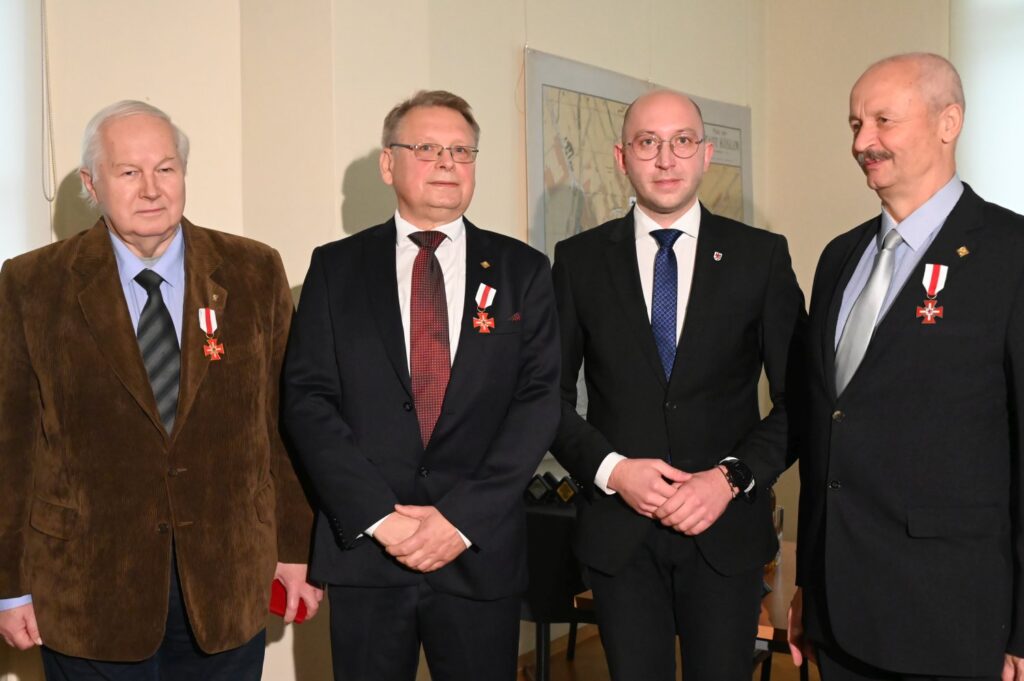 Czterech mężczyzn stoi obok siebie podczas uroczystego otwarcia wystawy; wszyscy są ubrani w eleganckie stroje, a trzej z nich mają przypięte do marynarek odznaczenia; w tle widoczna jest mapa powieszona na ścianie (kliknięcie powiększy zdjęcie).