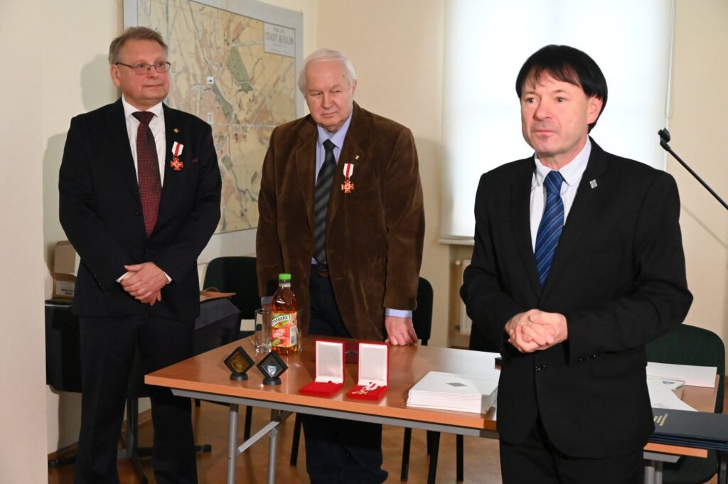 Trzech mężczyzn stoi przy stole z dokumentami, pudełkami z odznaczeniami i innymi materiałami związanymi z uroczystością; jeden z nich przemawia do zgromadzonych, a pozostali stoją obok i obserwują wydarzenie (kliknięcie powiększy zdjęcie).