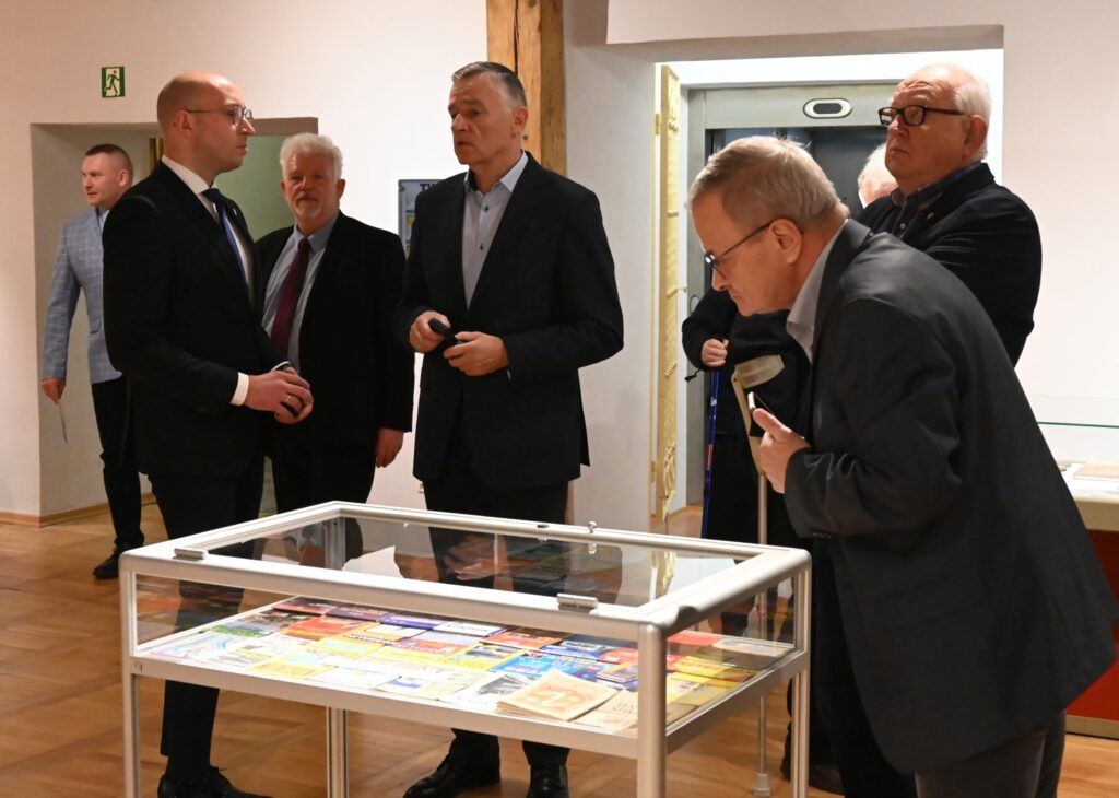 Grupa mężczyzn rozmawia w sali wystawowej przy przeszklonej gablocie; jeden z uczestników pochyla się nad eksponatami i uważnie je ogląda, podczas gdy pozostali obserwują prezentowane zbiory i prowadzą rozmowę (kliknięcie powiększy zdjęcie).