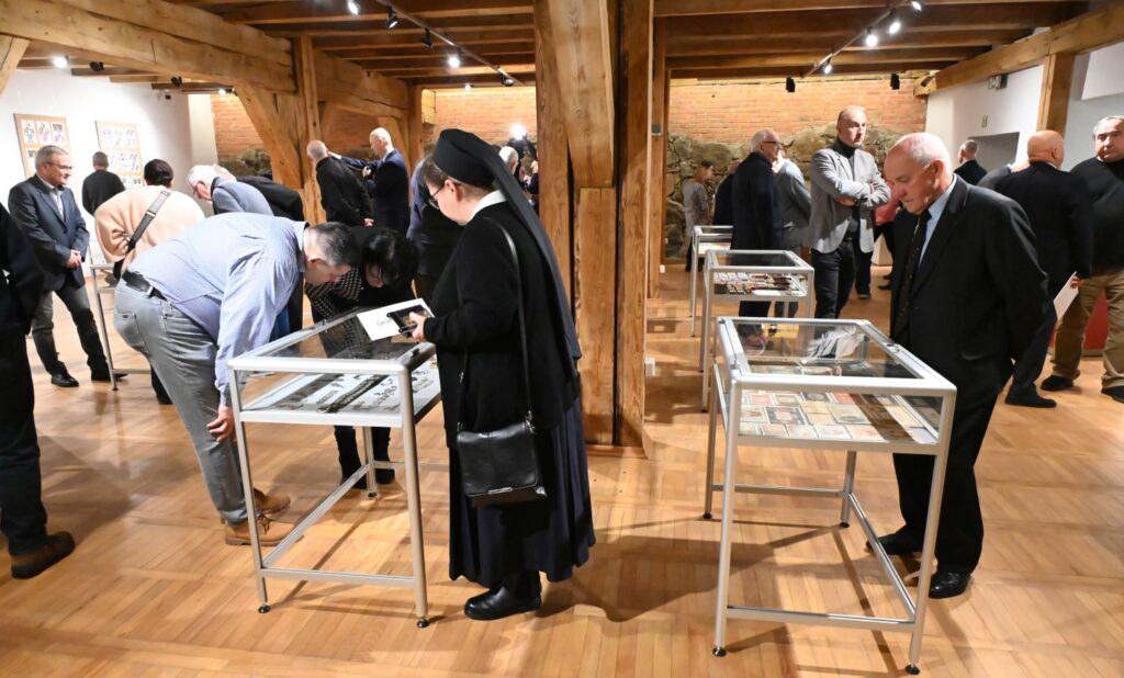Liczna grupa zwiedzających ogląda kilka przeszklonych gablot ustawionych w sali wystawowej; część osób pochyla się nad eksponatami, dokładnie przyglądając się prezentowanym zbiorom (kliknięcie powiększy zdjęcie).