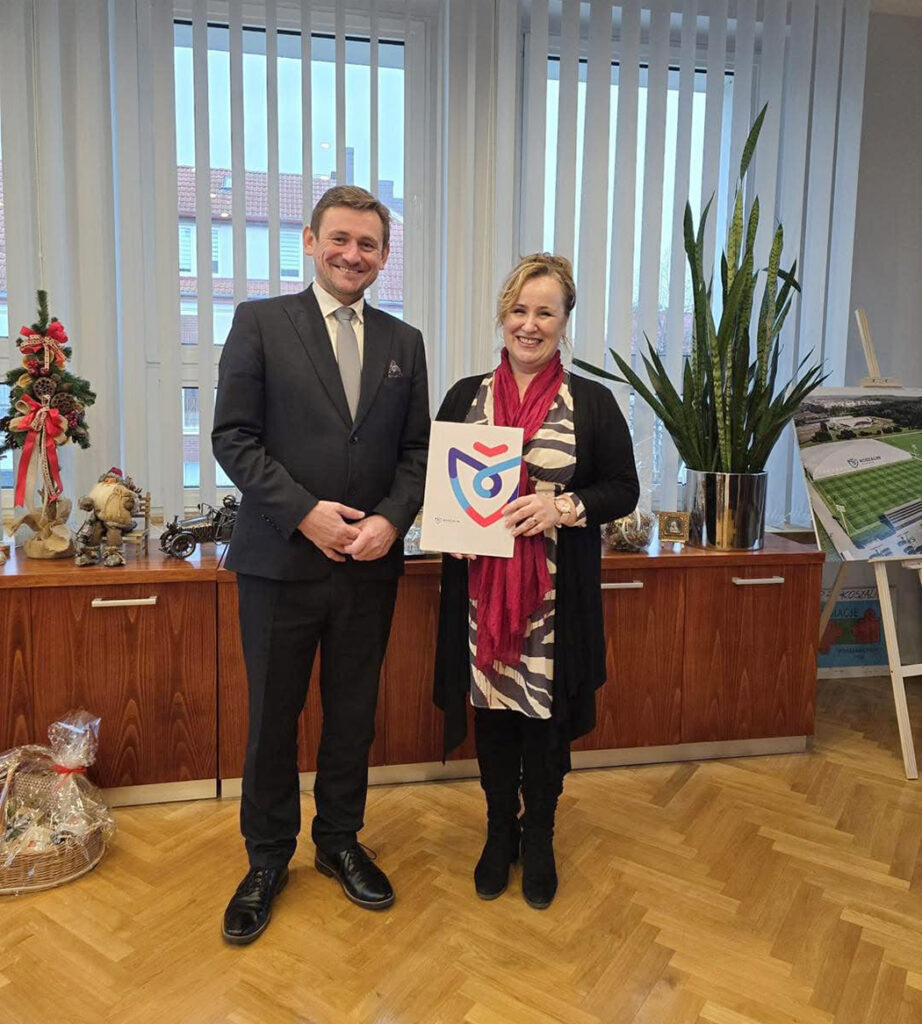 Kobieta trzyma dokument z kolorowym logo i stoi obok uśmiechniętego mężczyzny w garniturze, pozując do zdjęcia w biurze przy szafce z dekoracjami i rośliną (kliknięcie powiększy zdjęcie).