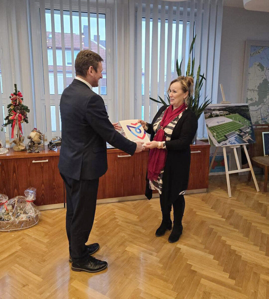 Mężczyzna w garniturze wręcza kobiecie dokument z kolorowym logo w biurze, w tle szafka z dekoracjami, roślina i plansza projektu (kliknięcie powiększy zdjęcie).