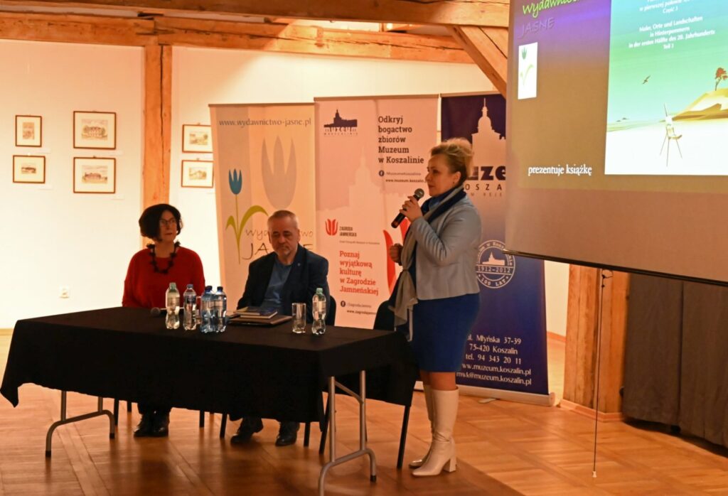 Przy stole siedzą uczestnicy wydarzenia, a stojąca obok kobieta z mikrofonem prowadzi rozmowę; w tle widoczne są materiały promocyjne muzeum i ekran z prezentacją (kliknięcie powiększy zdjęcie).