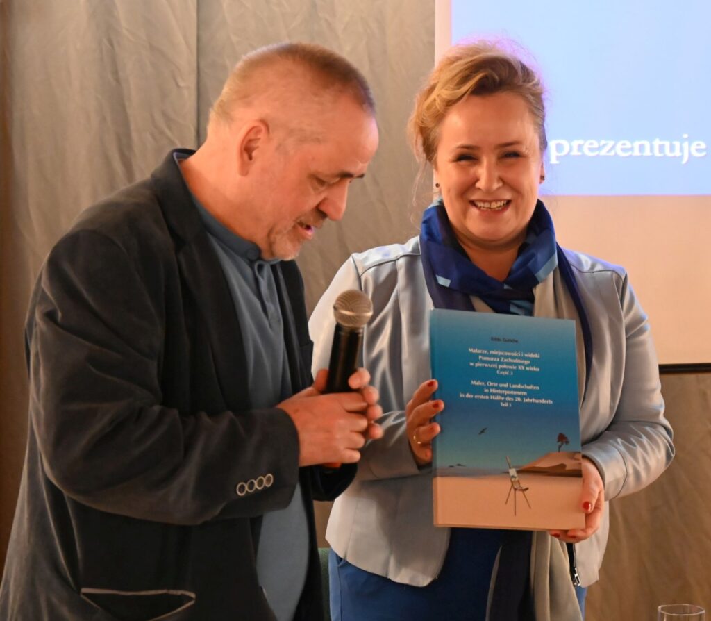 Mężczyzna i kobieta podczas spotkania promocyjnego; kobieta trzyma książkę z okładką przedstawiającą nadmorski pejzaż, a mężczyzna obok niej trzyma mikrofon; w tle widoczny jest ekran z prezentacją (kliknięcie powiększy zdjęcie).