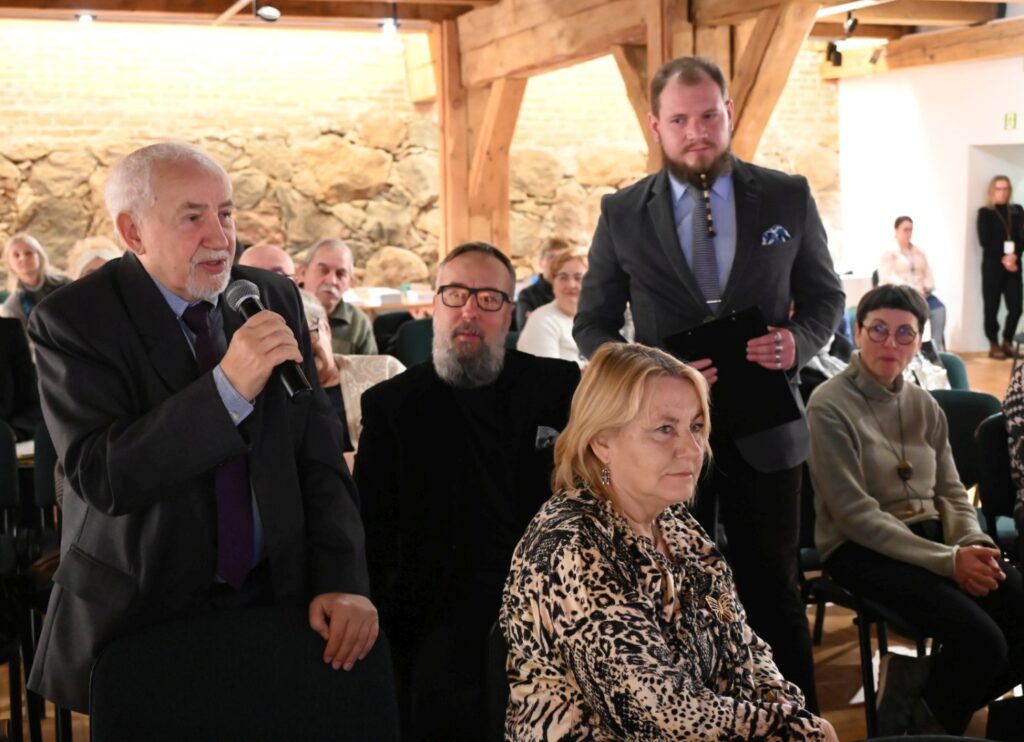 Uczestnik spotkania zabiera głos z miejsca na widowni, trzymając mikrofon; w pierwszym planie siedzi kobieta, a za nią zgromadzona publiczność, w tym osoby stojące przy ścianie (kliknięcie powiększy zdjęcie).