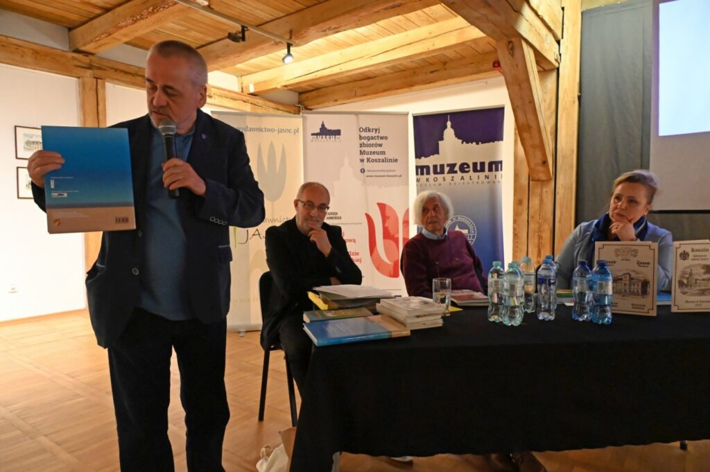 Mężczyzna przemawia przy stole podczas spotkania promocyjnego, trzymając mikrofon i książkę; przy stole siedzą pozostali uczestnicy, a w tle widoczne są materiały promocyjne i ekran prezentacji (kliknięcie powiększy zdjęcie).