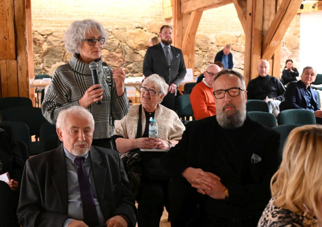 Spotkanie promocyjne podczas dyskusji; stojąca kobieta z mikrofonem zabiera głos, a w pierwszym planie siedzą uczestnicy wydarzenia; w tle pozostali słuchacze (kliknięcie powiększy zdjęcie).