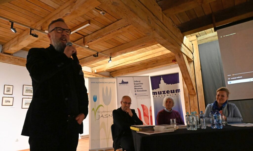 Mężczyzna w czarnym stroju przemawia do uczestników z mikrofonem; za nim przy stole siedzą osoby prowadzące spotkanie, a w tle widoczne są banery promocyjne i fragment ekspozycji muzealnej (kliknięcie powiększy zdjęcie).