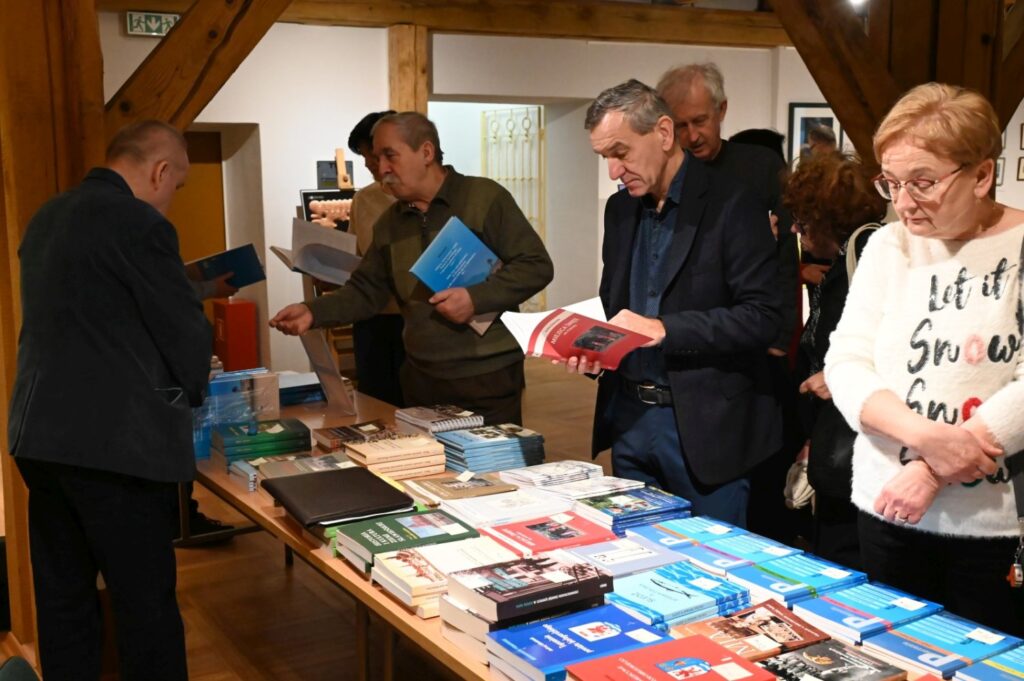 Stół z publikacjami towarzyszącymi spotkaniu; na pierwszym planie ułożono liczne książki i katalogi, a kilka osób przegląda wydawnictwa i rozmawia przy stole; w tle widoczna jest ekspozycja muzealna (kliknięcie powiększy zdjęcie).