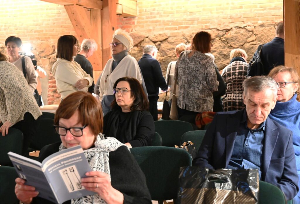 Uczestnicy spotkania rozmawiają i przemieszczają się po sali po zakończeniu części prezentacyjnej; część osób siedzi na krzesłach, a w tle widoczna jest ceglana ściana i drewniana konstrukcja wnętrza (kliknięcie powiększy zdjęcie).