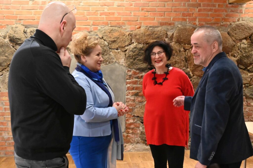Kilkoro uczestników spotkania rozmawia w kuluarach na tle ceglanej ściany; w grupie znajduje się kobieta w czerwonym swetrze oraz mężczyzna trzymający książkę, a obok nich stoi fotograf lub operator kamery (kliknięcie powiększy zdjęcie).