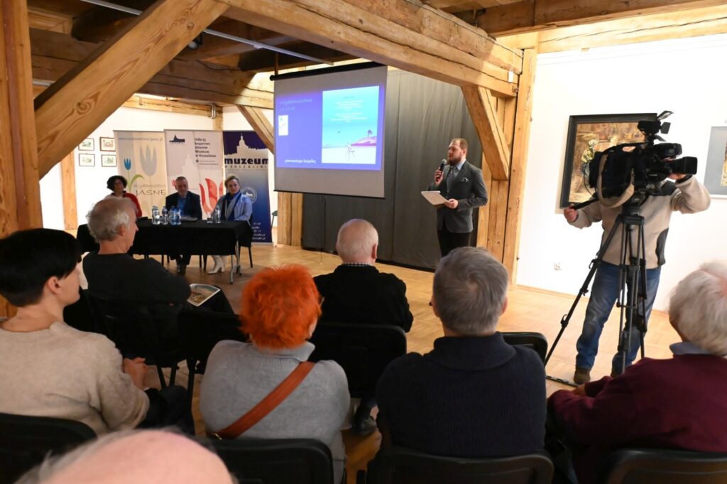 Spotkanie promocyjne w trakcie prezentacji; mężczyzna stoi przed publicznością z mikrofonem, a obok znajduje się kamera na statywie rejestrująca wydarzenie; za nimi widoczny jest ekran z prezentacją i stół prelegentów (kliknięcie powiększy zdjęcie).