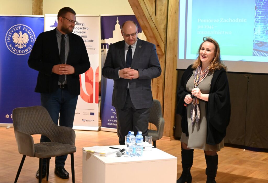 Trzy osoby stojące na scenie podczas spotkania konferencyjnego obok stolik z wodą i mikrofonami w tle banery instytucji (kliknięcie powiększy zdjęcie).