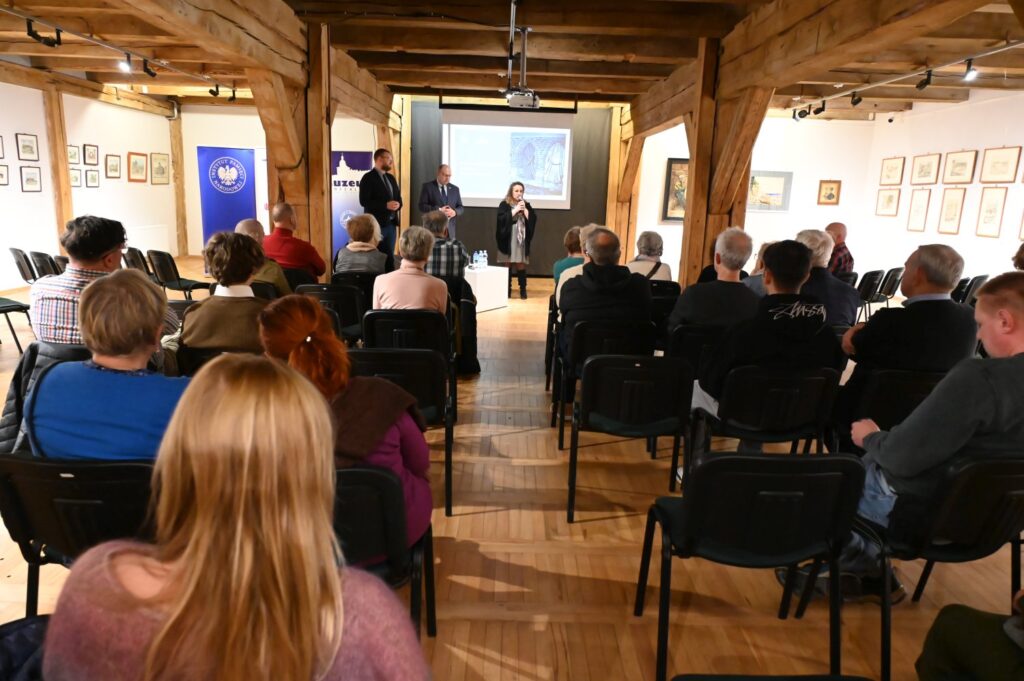 Uczestnicy konferencji siedzący na krzesłach w sali wystawowej słuchają wystąpienia prowadzonego na scenie (kliknięcie powiększy zdjęcie).