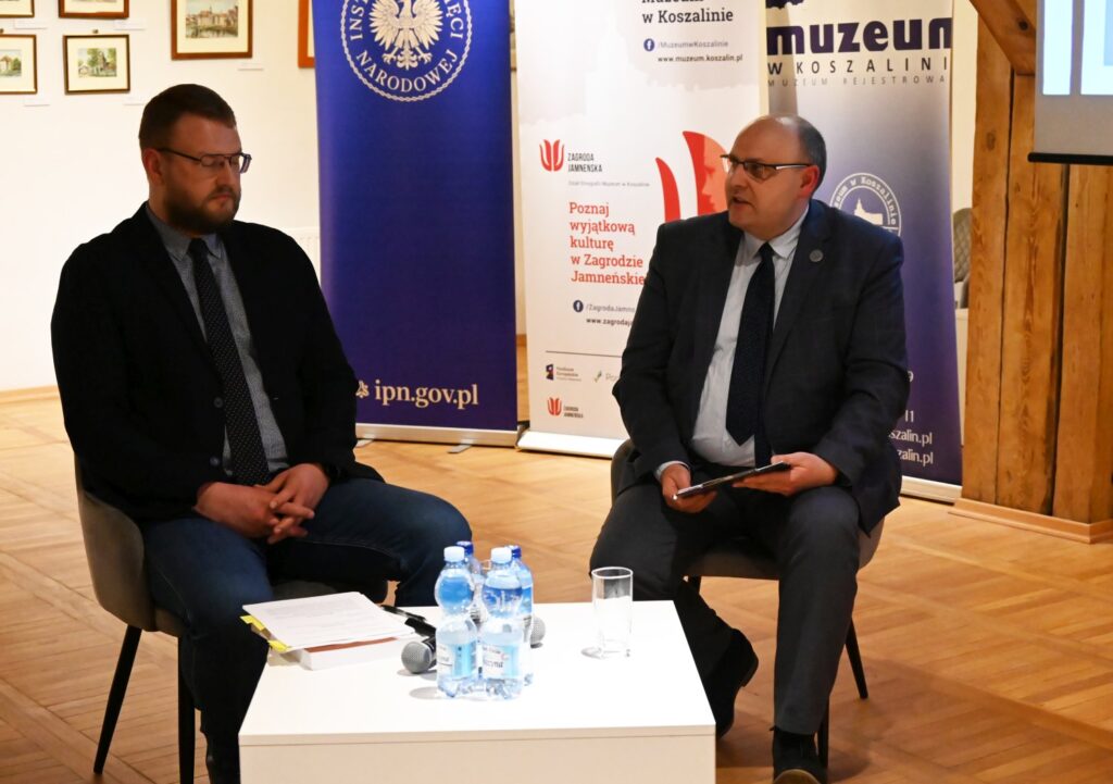 Dwóch mężczyzn siedzących na scenie podczas rozmowy konferencyjnej obok stolik z wodą i mikrofonami w tle banery instytucji (kliknięcie powiększy zdjęcie).