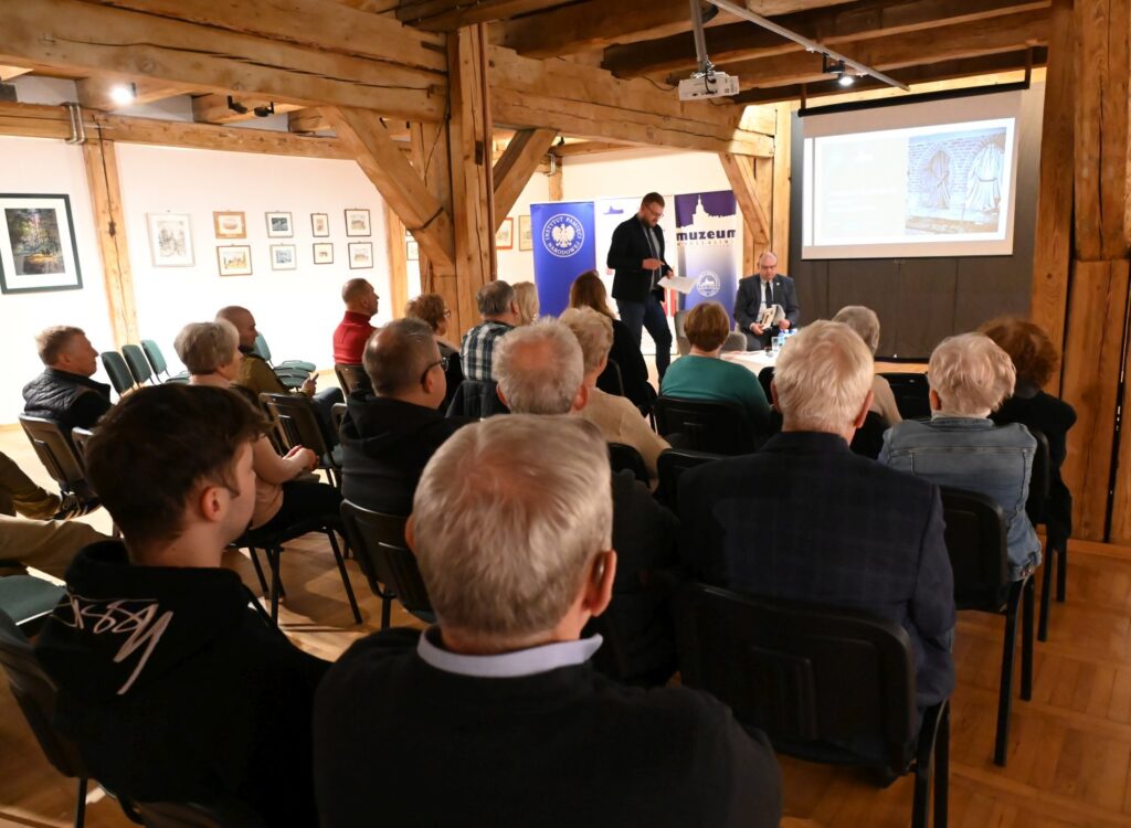 Widok sali wystawowej z tyłu gdzie uczestnicy siedzą na krzesłach i oglądają prezentację wyświetlaną na ekranie (kliknięcie powiększy zdjęcie).