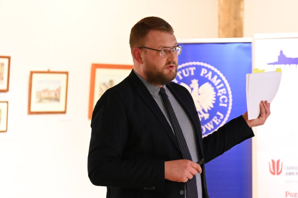 Mężczyzna w garniturze przemawia trzymając kartkę na tle baneru instytucji i plansz wystawowych (kliknięcie powiększy zdjęcie).
