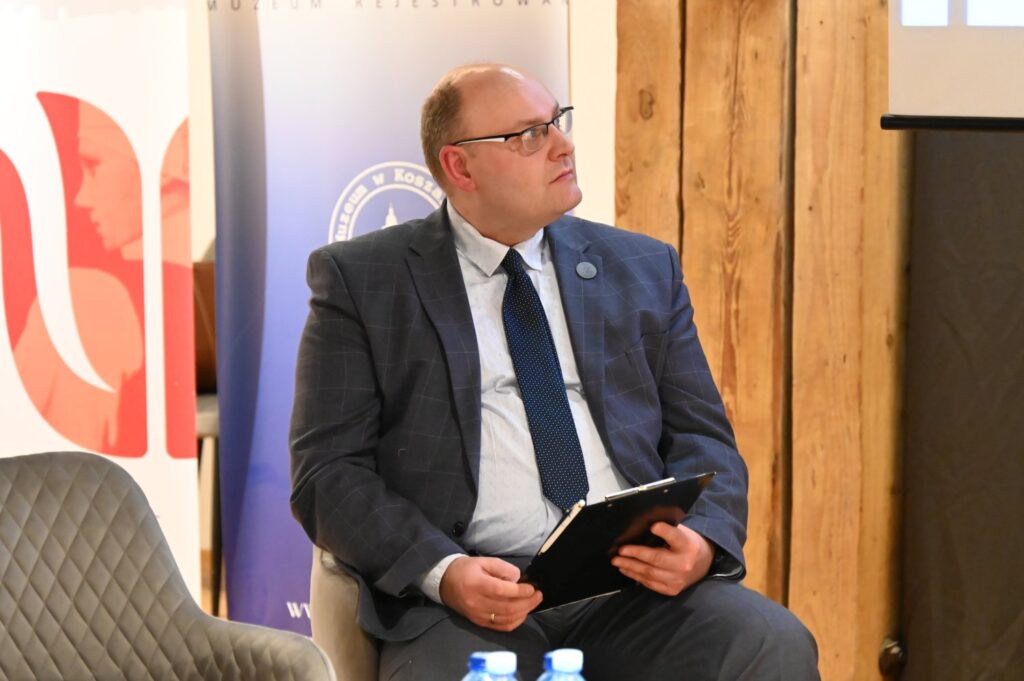 Prelegent siedzący na scenie z tabletem w rękach słucha dyskusji podczas konferencji (kliknięcie powiększy zdjęcie).