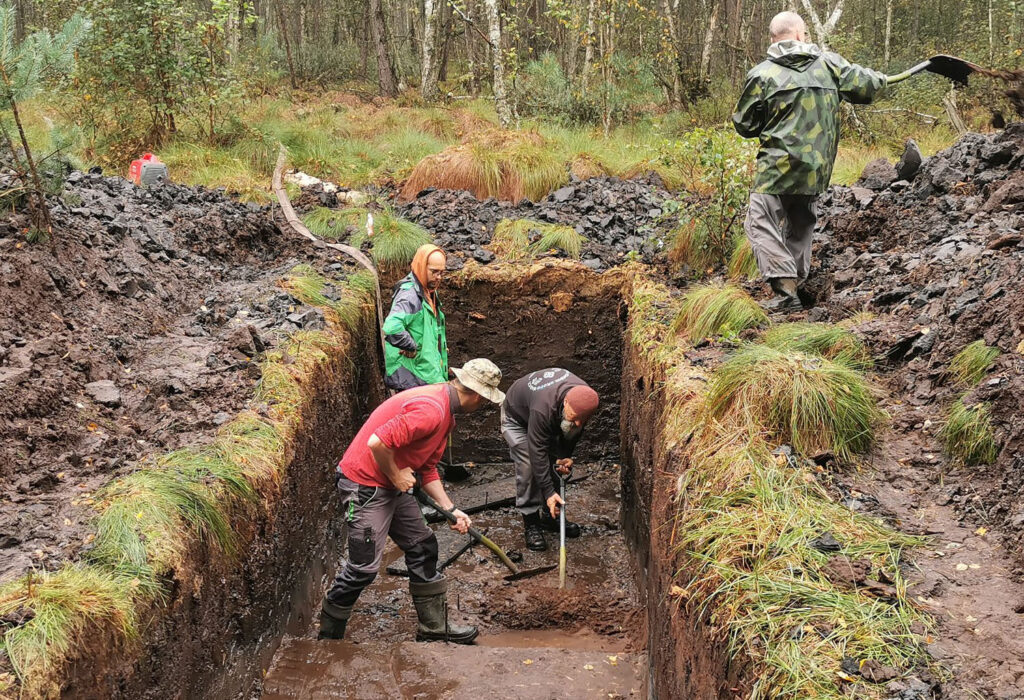 Archeolodzy prowadzą prace w głębokim wykopie, odkopując kolejne warstwy ziemi, jedna osoba stoi na krawędzi z łopatą, inni pracują na dnie wykopu w wodzie (kliknięcie powiększy zdjęcie).