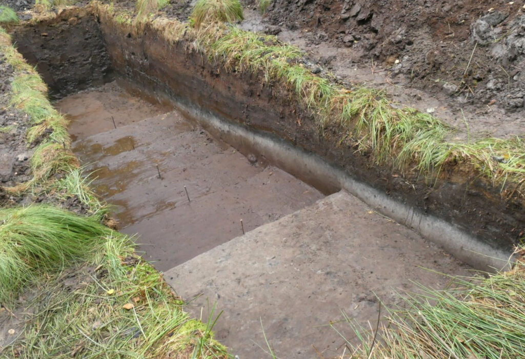 Oczyszczony profil wykopu archeologicznego ukazujący warstwy gleby oraz płaską powierzchnię dna z zaznaczonymi punktami pomiarowymi (kliknięcie powiększy zdjęcie).