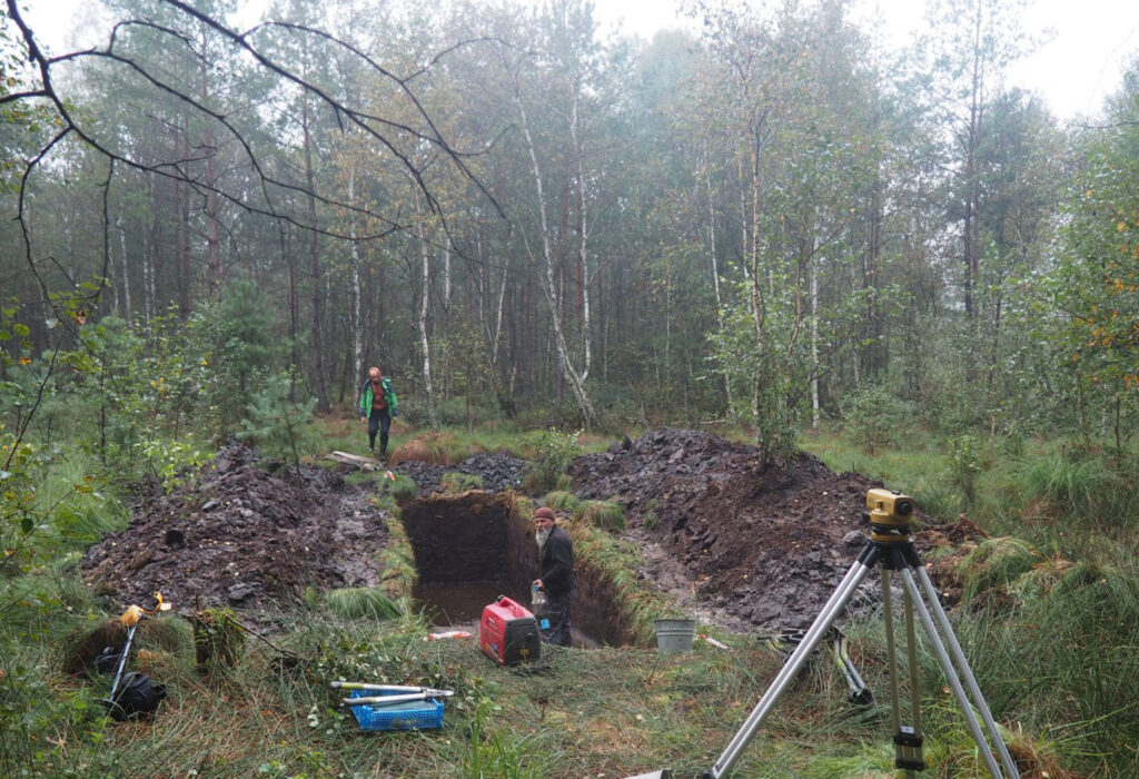 Szeroki widok stanowiska archeologicznego w lesie z głębokim wykopem, sprzętem pomiarowym na statywie oraz osobami pracującymi i nadzorującymi badania (kliknięcie powiększy zdjęcie).