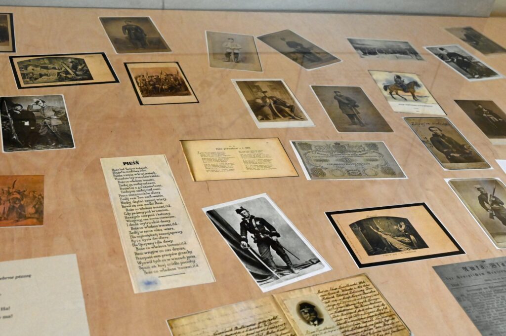 Zestaw materiałów związanych z powstaniem styczniowym, obejmujący fotografie, dokumenty, pamiątki osobiste, druki i ilustracje; materiały zostały rozmieszczone na płaskiej powierzchni ekspozycyjnej i tworzą przekrojowy obraz wydarzeń oraz osób związanych z powstaniem (kliknięcie powiększy zdjęcie).