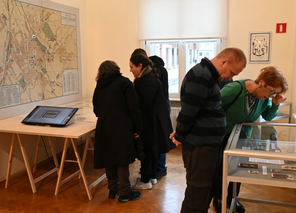 Zwiedzający oglądają ekspozycję, kilka osób pochyla się nad gablotami z prezentowanymi materiałami, a na ścianie widoczna jest mapa; w sali znajduje się również stanowisko multimedialne uzupełniające wystawę (kliknięcie powiększy zdjęcie).