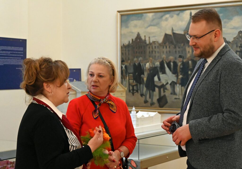 Rozmowa uczestników wydarzenia w przestrzeni wystawy, kilka osób rozmawia przy muzealnych gablotach i prezentowanych dziełach; w tle widoczny jest obraz oraz fragment ekspozycji (kliknięcie powiększy zdjęcie).