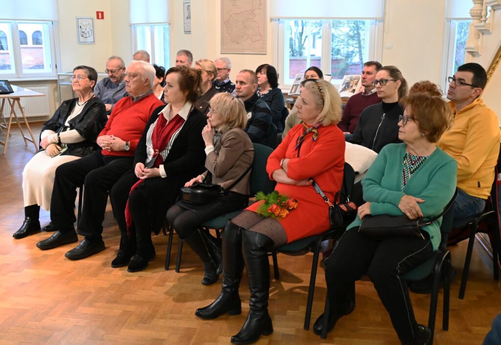 Publiczność zgromadzona podczas wydarzenia w muzeum, uczestnicy siedzą w rzędach i słuchają prowadzących; wśród obecnych widoczne są osoby w różnym wieku, a w tle znajdują się gabloty i elementy wystawy (kliknięcie powiększy zdjęcie).