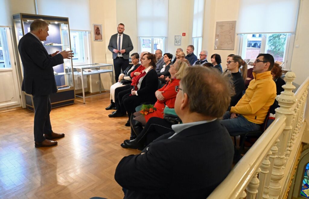 Publiczność podczas spotkania towarzyszącego otwarciu wystawy, uczestnicy siedzą naprzeciw osób prowadzących wydarzenie; w sali widoczne są gabloty z prezentowanymi obiektami oraz fragmenty ekspozycji (kliknięcie powiększy zdjęcie).