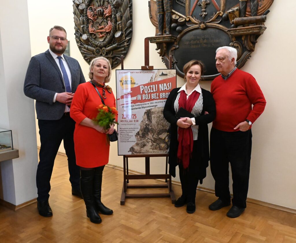 Grupa osób uczestniczących w otwarciu wystawy, stojących przy planszy informującej o wydarzeniu i prezentującej jego temat; jedna z osób trzyma bukiet kwiatów, a w tle widoczne są muzealne eksponaty i elementy dekoracyjne wnętrza (kliknięcie powiększy zdjęcie).