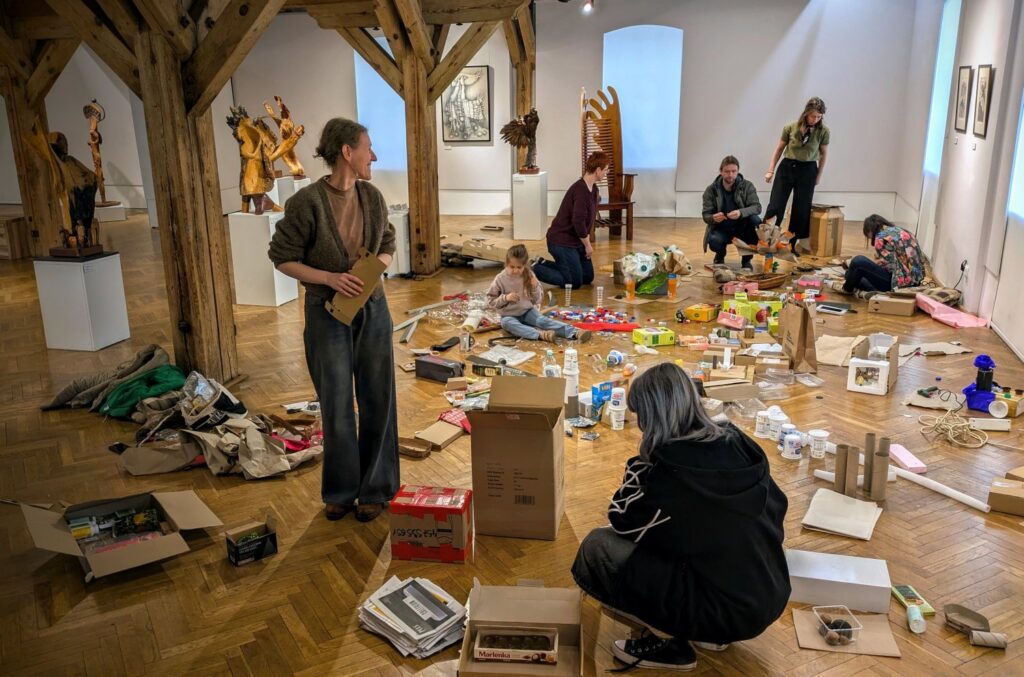 Uczestnicy podczas warsztatów artystycznych, osoby tworzą własne prace z wykorzystaniem kartonów, papieru, opakowań i innych materiałów; zajęcia odbywają się w przestrzeni wystawy, wśród prezentowanych rzeźb i instalacji (kliknięcie powiększy zdjęcie).