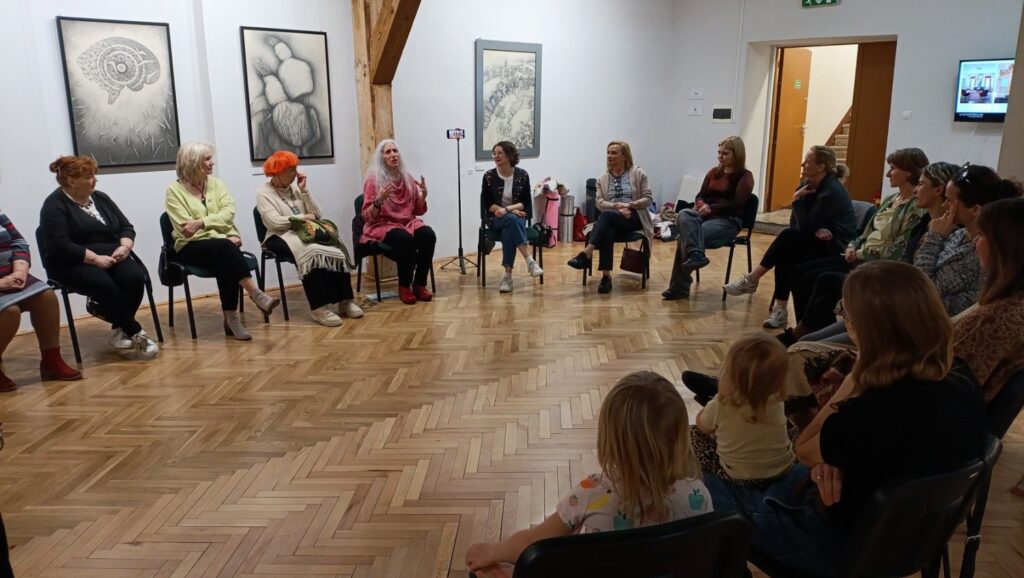Grupa osób siedzi w kręgu i uczestniczy w rozmowie lub zajęciach; wokół znajdują się prezentowane prace artystyczne oraz elementy ekspozycji (kliknięcie powiększy zdjęcie).