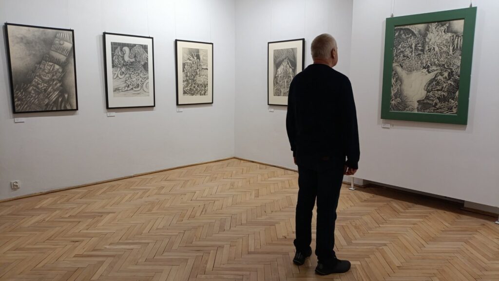 Mężczyzna ogląda grafiki prezentowane na ścianie sali wystawowej; w przestrzeni znajduje się kilka oprawionych prac o zróżnicowanej tematyce i formie, tworzących część ekspozycji (kliknięcie powiększy zdjęcie).