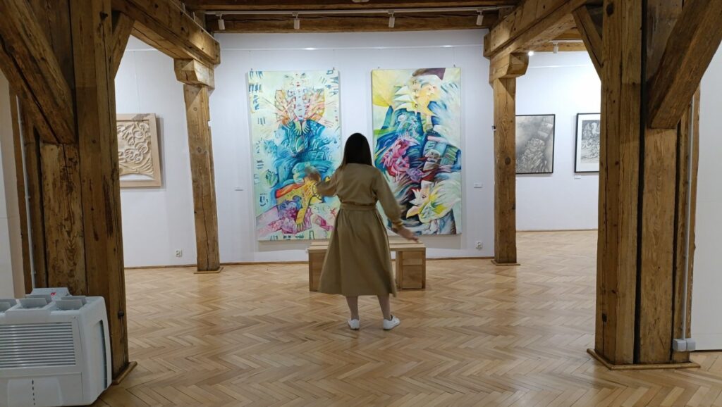 Kobieta ogląda wielkoformatowe, kolorowe prace prezentowane na ścianie galerii; w sali widoczne są również inne grafiki oraz elementy wystawy rozmieszczone w przestrzeni muzeum (kliknięcie powiększy zdjęcie).