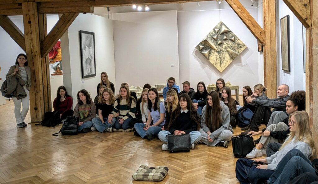 Grupa młodzieży uczestniczy w zajęciach edukacyjnych w przestrzeni wystawy; uczestnicy siedzą na podłodze i słuchają prowadzącej, a wokół nich prezentowane są prace artystyczne (kliknięcie powiększy zdjęcie).