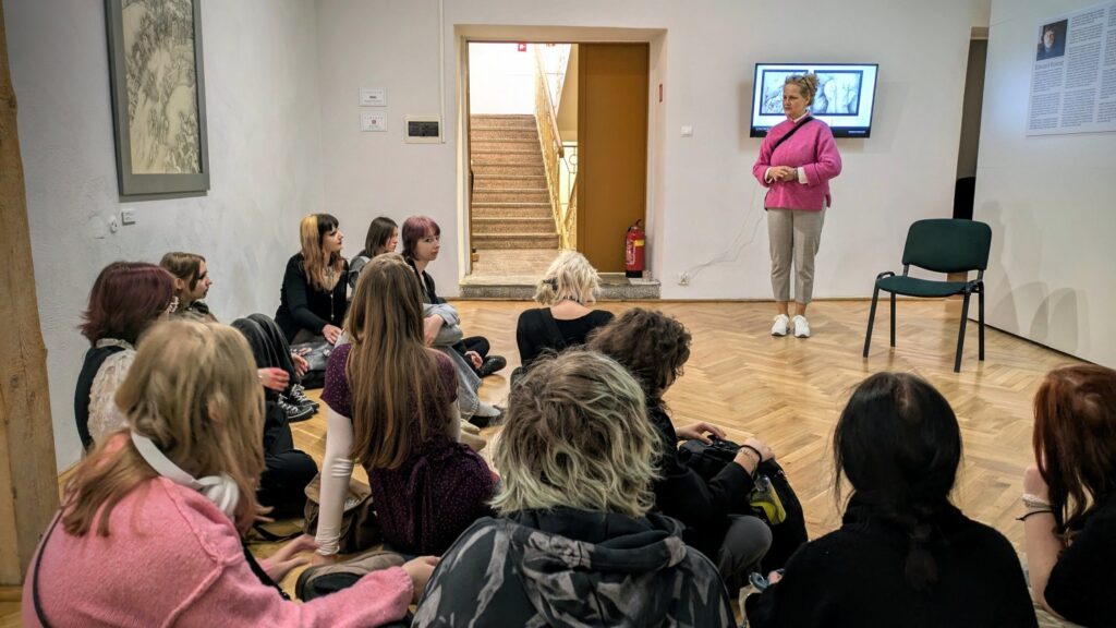 Uczestnicy zajęć siedzą na podłodze w sali wystawowej i słuchają prowadzącej stojącej przy ścianie; spotkanie odbywa się w bezpośrednim otoczeniu prezentowanych prac (kliknięcie powiększy zdjęcie).