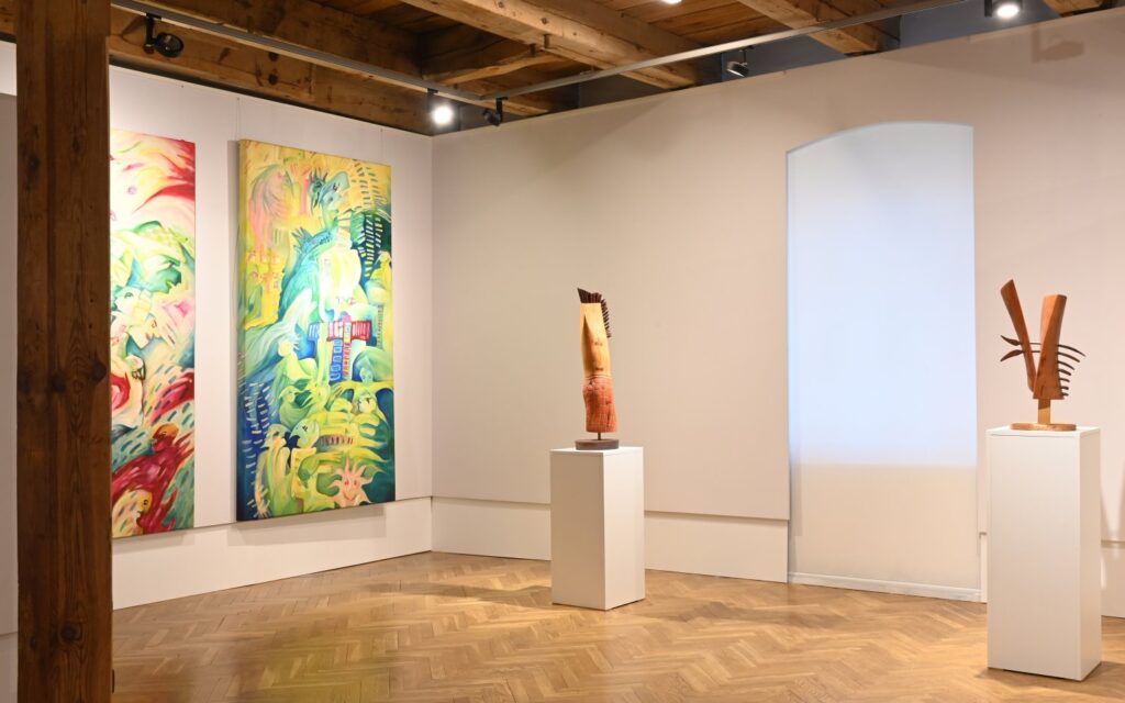 Fragment ekspozycji z kolorowymi obrazami oraz niewielkimi rzeźbami ustawionymi na postumentach; prace rozmieszczono w otwartej przestrzeni galerii, której wnętrze tworzą drewniane belki i jasne ściany (kliknięcie powiększy zdjęcie).