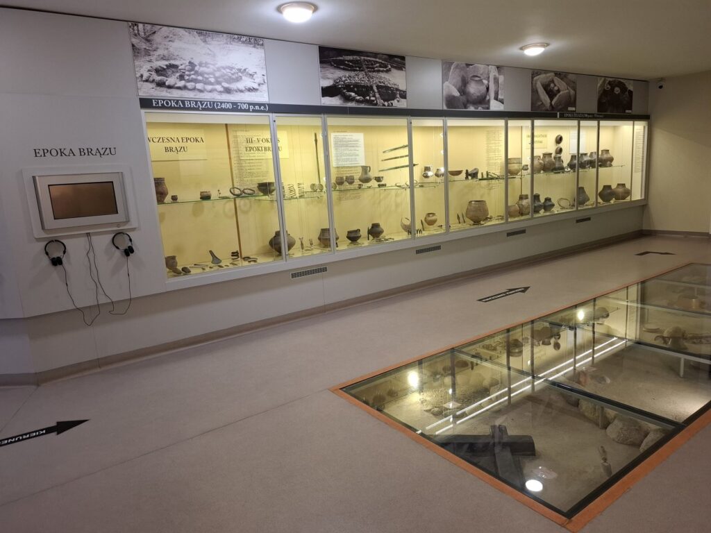 Sala wystawy archeologicznej z gablotami ściennymi oraz przeszkloną ekspozycją podłogową, prezentacja znalezisk z różnych epok (kliknięcie powiększy zdjęcie)