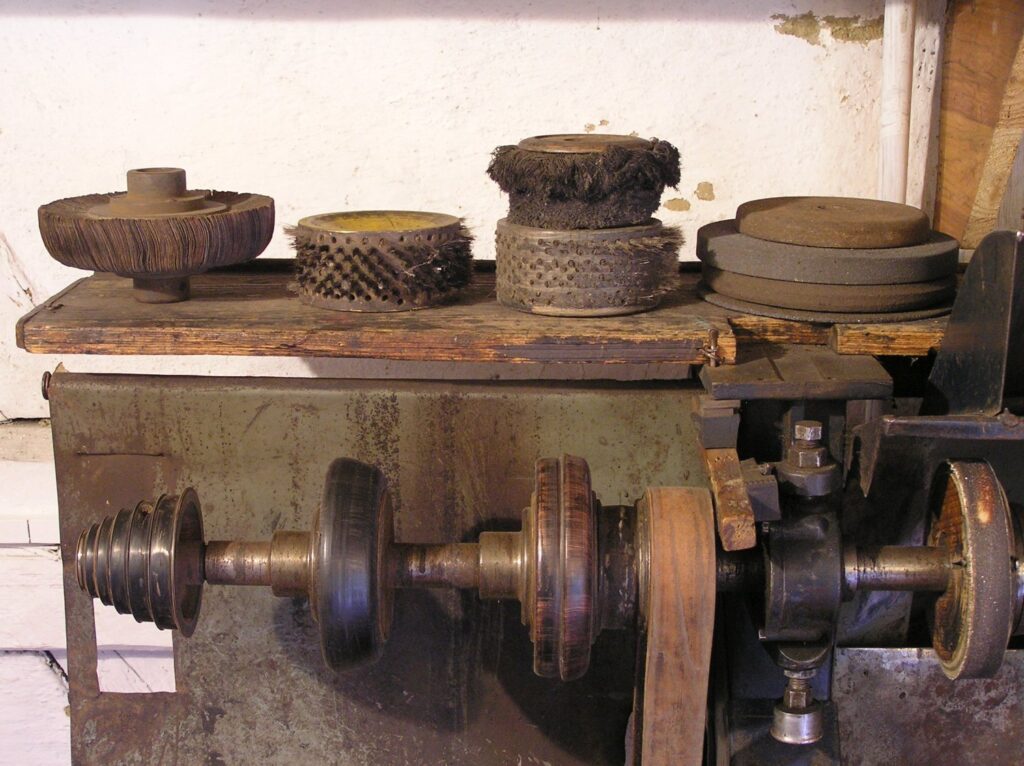 Zabytkowa maszyna do obróbki obuwia z metalowym mechanizmem oraz tarczami roboczymi, ustawiona we wnętrzu warsztatu szewskiego (kliknięcie powiększy zdjęcie).
