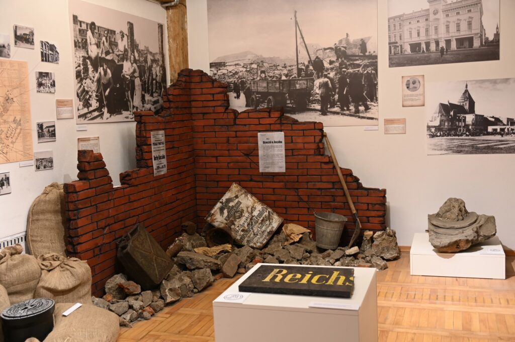 Aranżacja ekspozycyjna z fragmentem zburzonego muru, gruzem i workami, na tle archiwalnych fotografii zniszczonego miasta (kliknięcie powiększy zdjęcie).