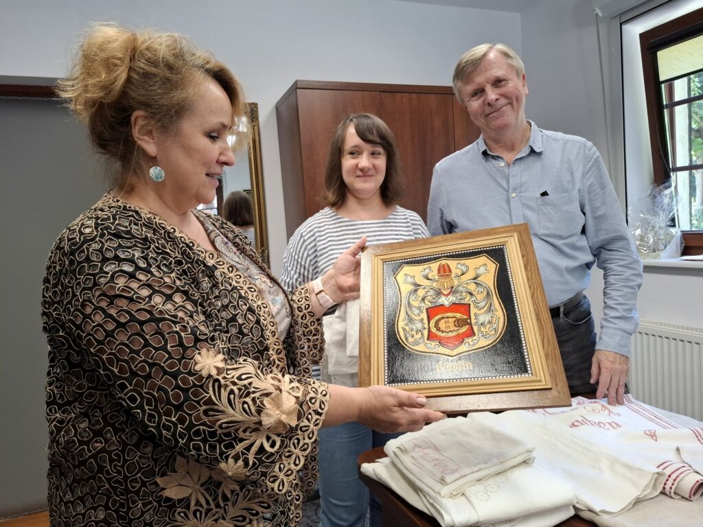 Trzy osoby prezentują artefakty przekazane do muzeum. Na pierwszym planie dyrektor pokazuje osadzony w ramce herb (kliknięcie spowoduje powiększenie zdjęcia).