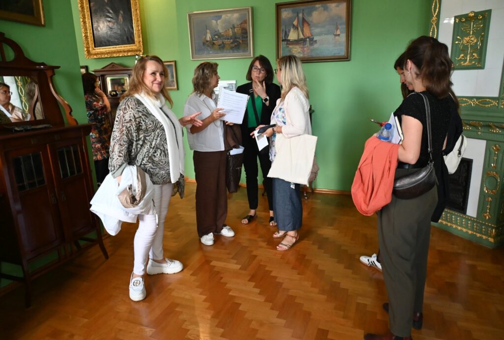 Członkowie francuskiej delegacji rozmawiają w zabytkowym wnętrzu muzeum z zielonymi ścianami i obrazami, trzymając materiały informacyjne (kliknięcie powiększy zdjęcie).