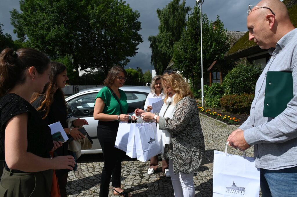 Uczestnicy wizyty spotykają się na zewnątrz muzeum, wymieniając się materiałami promocyjnymi i torbami z upominkami na tle zaparkowanego samochodu i zieleni (kliknięcie powiększy zdjęcie).